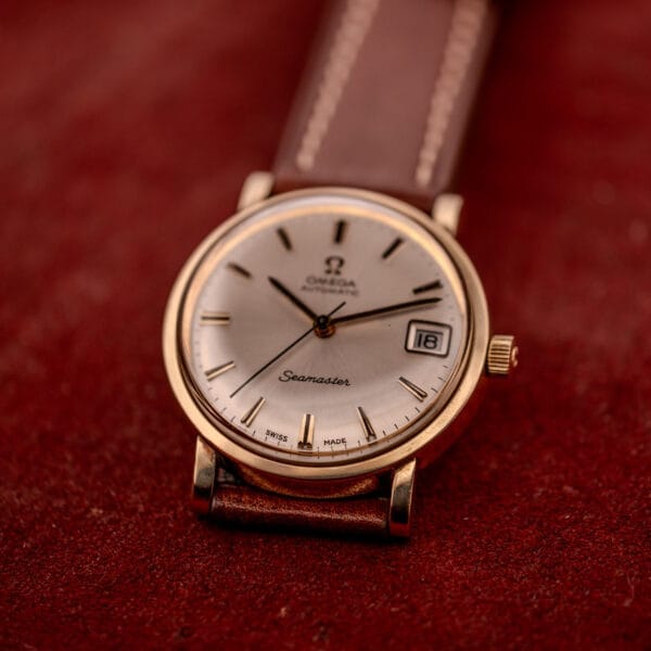 An Omega Automatic Seamaster Date Ref. KM 6323 with Cal. 563 movement and a brown leather strap rests on a red textured surface.