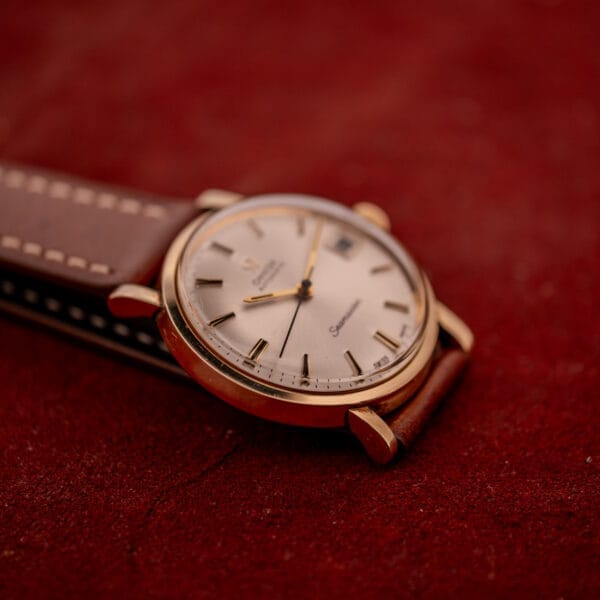 An Omega Automatic Seamaster Date Ref. KM 6323 Cal. 563 with a gold case and brown leather strap rests elegantly on a textured red surface.
