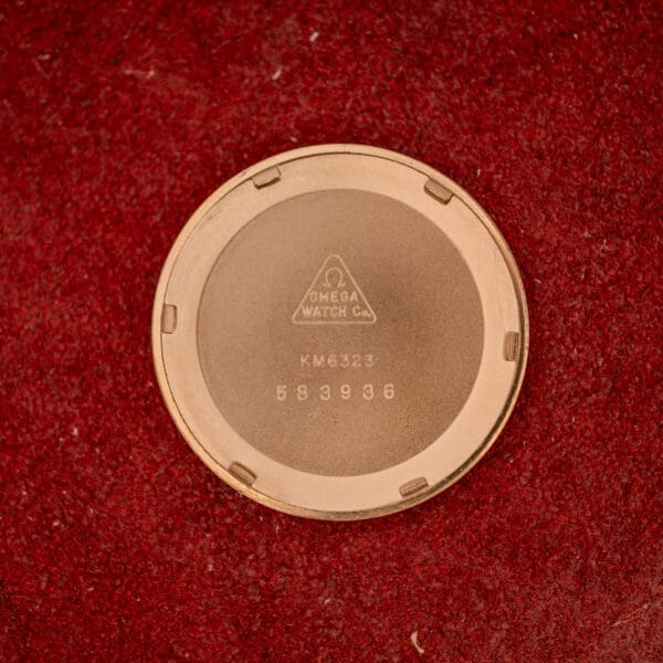 Case back of the Omega Automatic Seamaster Date Ref. KM 6323 Cal. 563, featuring numbers and KM 6323, displayed on a textured red surface.
