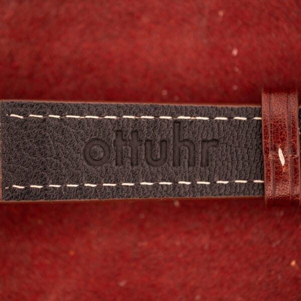 A close-up of a brown leather strap embossed with "ottuhr" on a red textured background, echoing the refined details of the Omega Automatic Seamster Date Ref. KM 6323 Cal. 563.