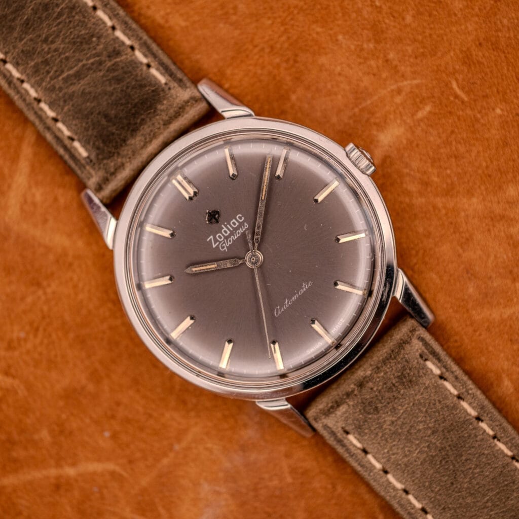 A Zodiac Glorious Automatic Ref. 702-917 Cal. 70-72 wristwatch with a brown leather strap rests on tan leather, highlighting its timeless design and robust stainless steel case.