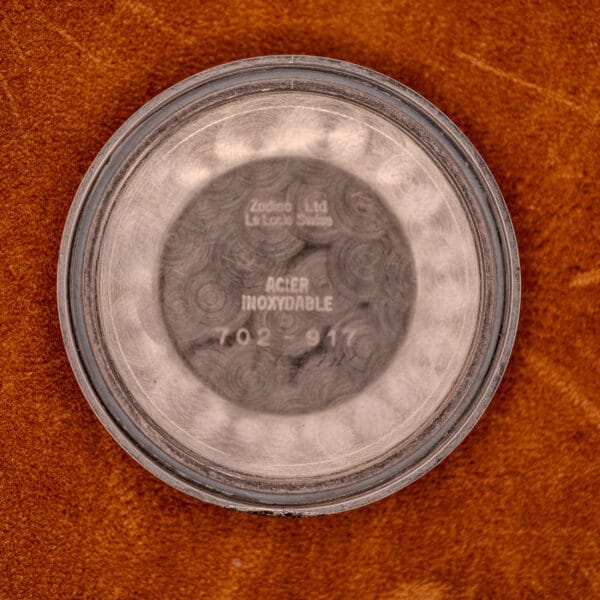 The Zodiac Glorious Automatic Ref. 702-917 Stainless Steel Cal. 70-72 watch case back, engraved with model details, rests on a brown textured surface and is viewed from above.