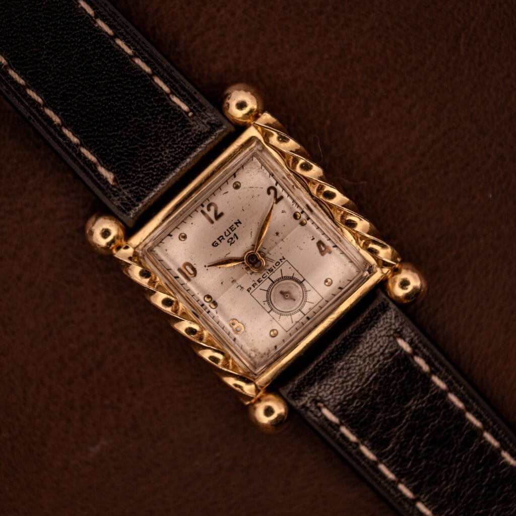 Gruen 21 Precision Art Deco Twisted Rope Case Ref. 335/530 wristwatch with a white dial and black leather strap, displayed on a brown background.