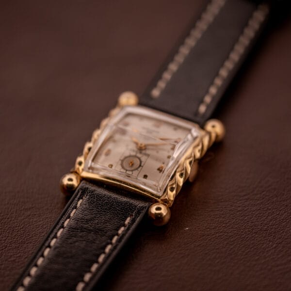Close-up of a Gruen 21 Precision Art Deco Twisted Rope Case Ref. 335 / 530 gold wristwatch with a black leather strap, resting on a brown surface.