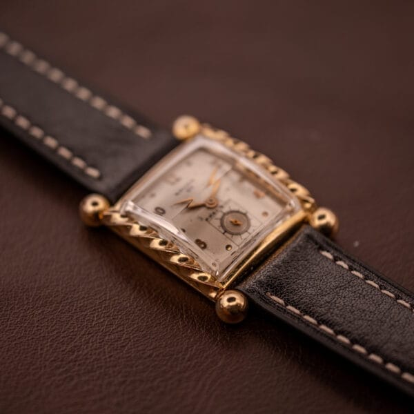 A Gruen 21 Precision Art Deco Twisted Rope Case Ref. 335 / 530 gold rectangular wristwatch with a black leather strap rests on a brown surface.