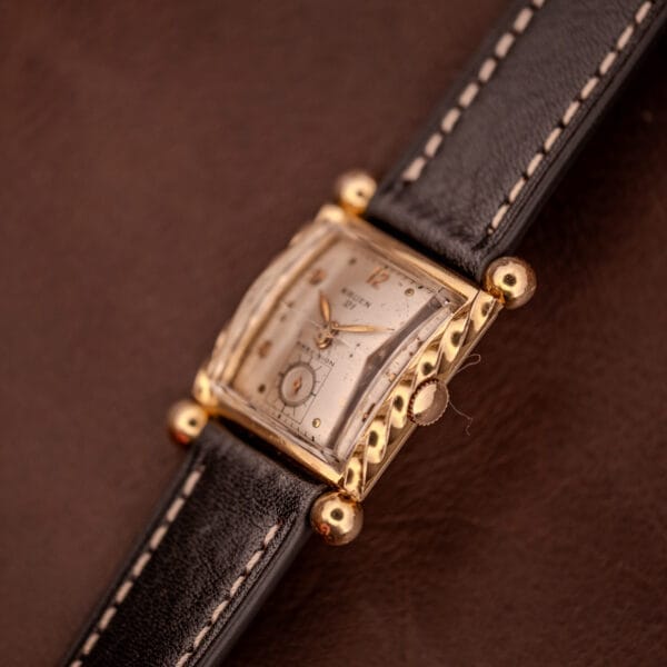 Gruen 21 Precision Art Deco Twisted Rope Case Ref. 335/530 vintage wristwatch with a gold square case and black leather strap displayed on a brown surface.