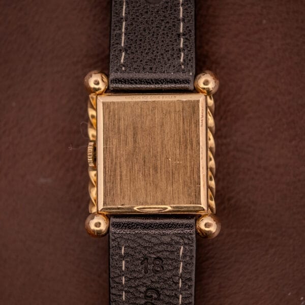 A square gold Gruen 21 Precision Art Deco Twisted Rope Case Ref. 335/530 wristwatch with a black leather strap, shown from the back against a brown background.