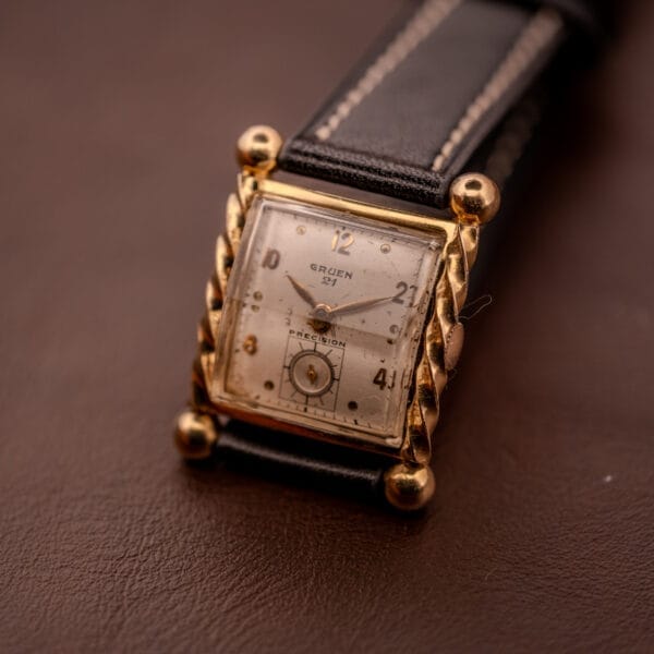 Gruen 21 Precision Art Deco Twisted Rope Case Ref. 335 / 530 wristwatch featuring a square gold case and black leather strap, elegantly displayed on a brown surface.