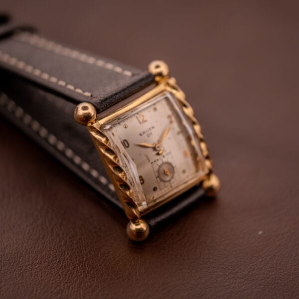 The Gruen 21 Precision Art Deco Twisted Rope Case Ref. 335 / 530 gold wristwatch features a square face and black leather strap, displayed on a brown surface.