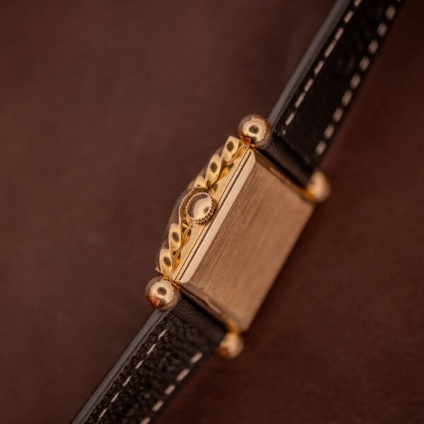 Close-up of a Gruen 21 Precision Art Deco Twisted Rope Case Ref. 335/530 watch with gold case, black leather strap, and twisted rope detail, viewed from the side against a brown background.