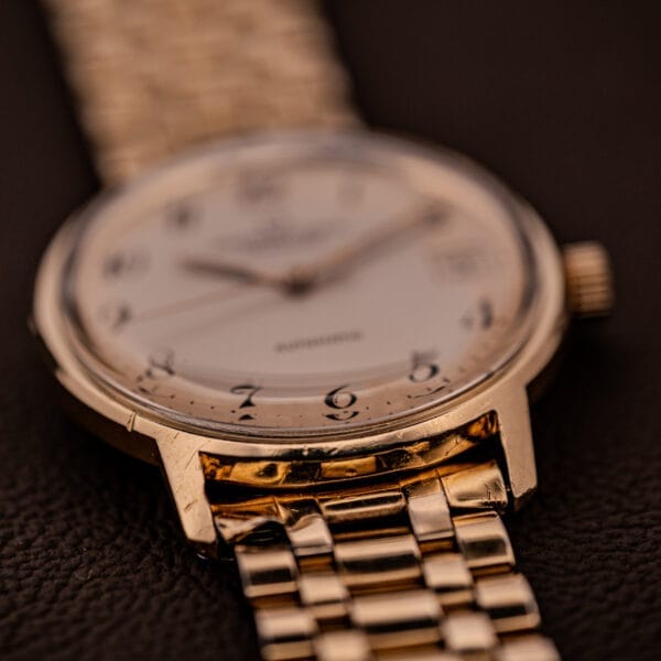 Close-up of a Universal Geneve Polerouter Date Arabic Numerals Dial Automatic Cal 215 gold wristwatch with metal band and white dial featuring black Arabic numerals, showcased on a dark surface.