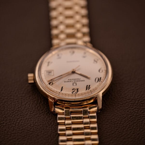 A Universal Geneve Polerouter Date Automatic Cal 215 wristwatch with Arabic numerals dial and a metal link band rests on a brown textured surface.