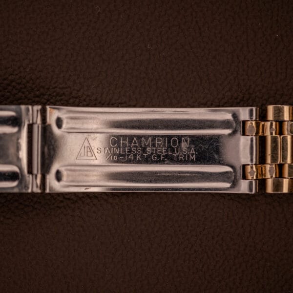 Close-up of a metal watch band clasp engraved with CHAMPION and material info, set on brown—an elegant detail for collectors of the Universal Geneve Polerouter Date Arabic Numerals Dial Automatic Cal 215.
