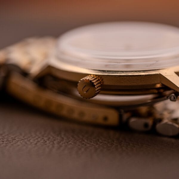 Close-up of a gold Universal Geneve Polerouter Date Arabic Numerals Dial Automatic Cal 215 watch crown and case, resting on brown leather.