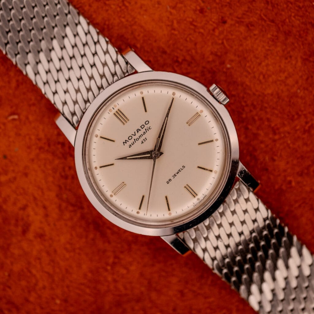 The Movado Automatic 431 Ref. 11201 features a stainless steel case and bracelet, shown on a textured orange surface.
