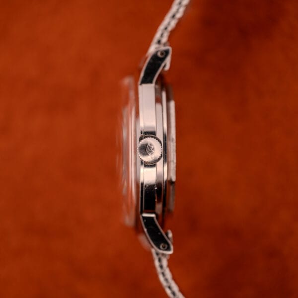 Side view of the Movado Automatic 431 Ref. 11201 with stainless steel case and bracelet, featuring a crown, displayed against a brown textured background.