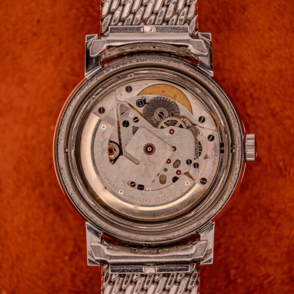 A close-up of the Movado Automatic 431 Ref. 11201 wristwatch, featuring a stainless steel case and bracelet, with a visible mechanical movement on an orange background.