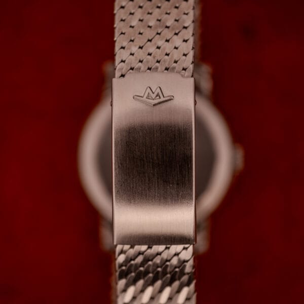Close-up of a stainless steel watch clasp with an engraved M logo, set against a blurred red background—an elegant detail from the Movado Automatic 431 Ref. 11201 Stainless Steel Case & Bracelet.