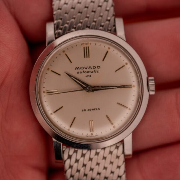 A close-up of a Movado Automatic 431 Ref. 11201 wristwatch, featuring a stainless steel case and bracelet with a cream dial.