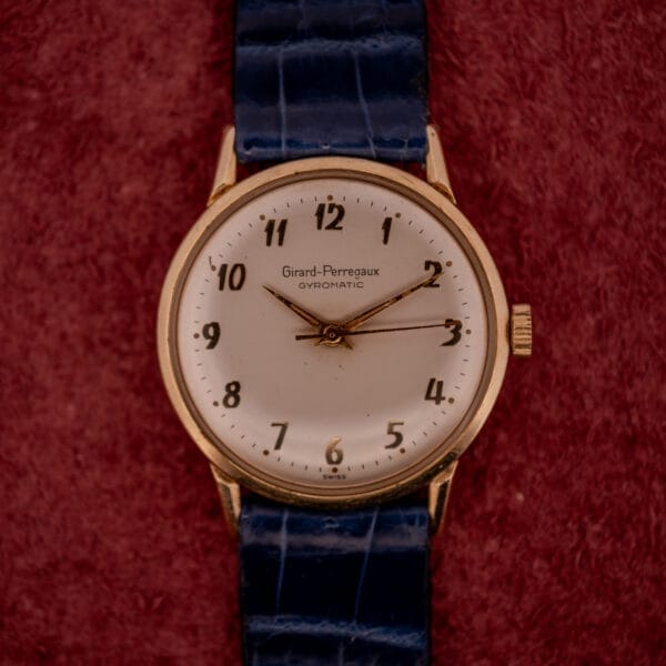 A Girard Perregaux Gyromatic with a white dial, gold Arabic numerals, and a blue strap sits on a red textured background.