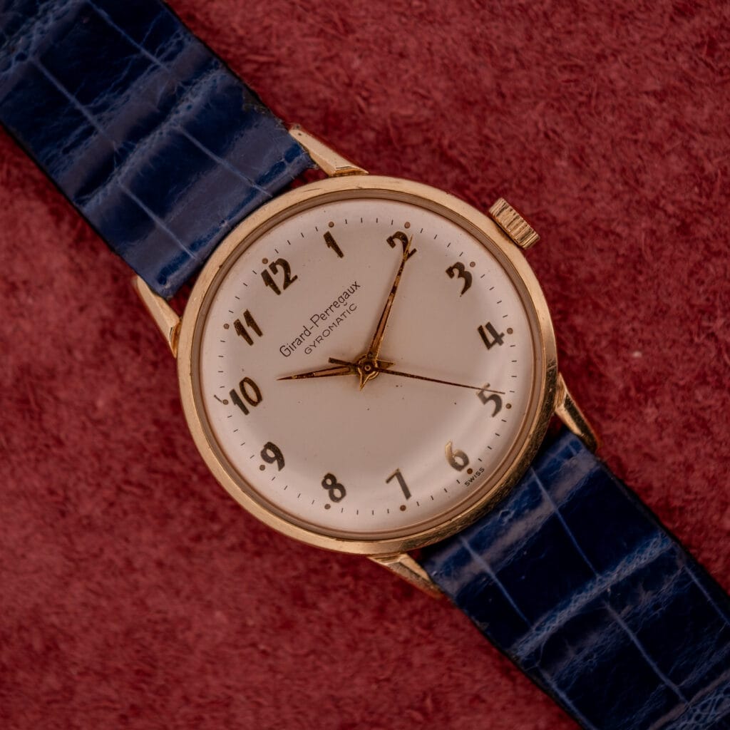 The Girard Perregaux Gyromatic White Dial & Gold Arabic Numerals watch rests elegantly on a textured red surface.