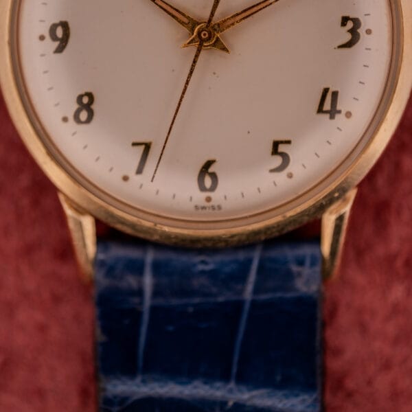 Close-up of a vintage Girard Perregaux Gyromatic with gold casing, white dial, and gold Arabic numerals, blue leather strap, displaying time near 6:00.