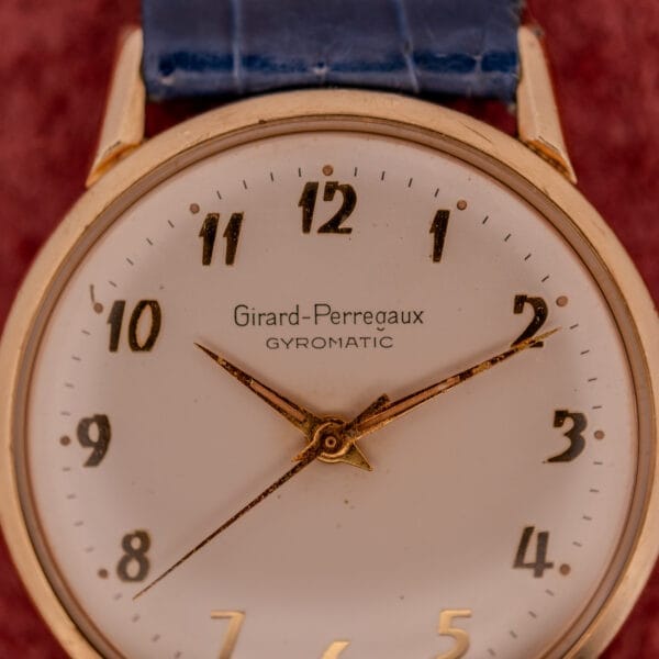 Girard Perregaux Gyromatic wristwatch featuring a white dial with gold Arabic numerals and a blue leather strap, displayed at 10:10.