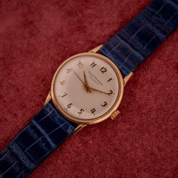 The Girard Perregaux Gyromatic White Dial & Gold Arabic Numerals wristwatch with a blue leather strap is displayed on a red textured surface, showing 10:09.