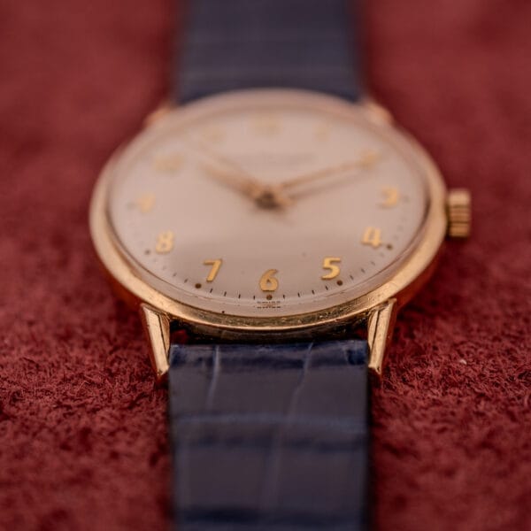 A Girard Perregaux Gyromatic with a white dial and gold Arabic numerals is elegantly displayed on a textured red surface.