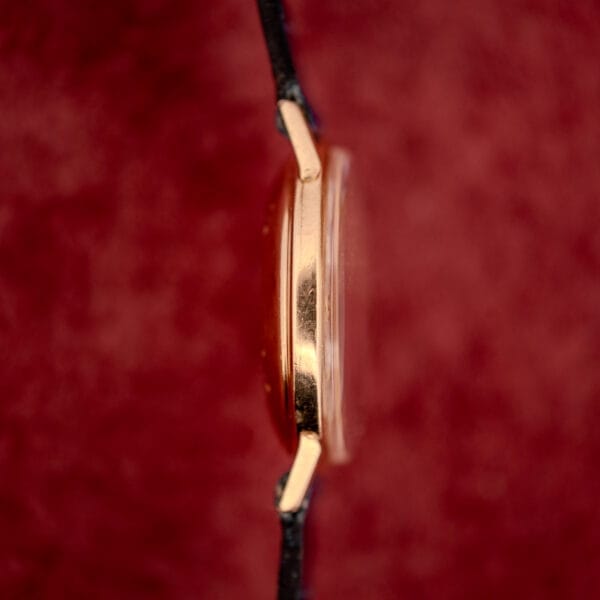 Side view of a thin Girard Perregaux Gyromatic White Dial & Gold Arabic Numerals watch featuring a gold case, black strap, and white dial, set against a blurred red background.