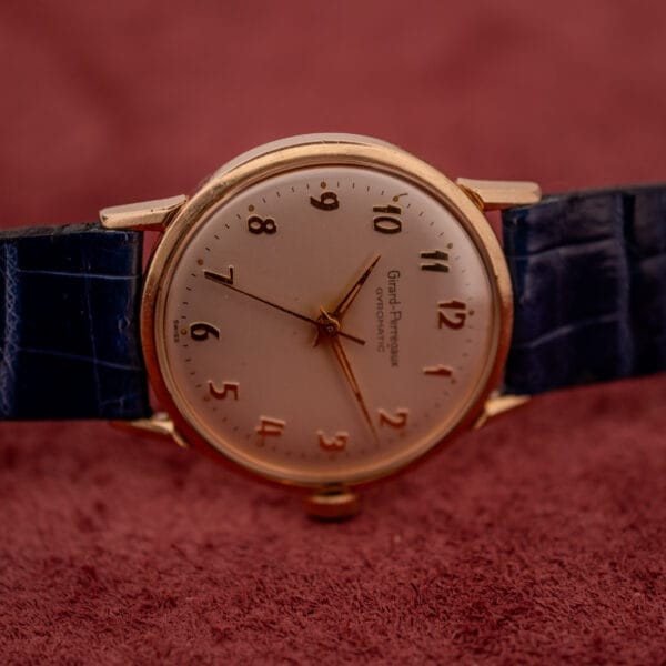 A Girard Perregaux Gyromatic with a white dial and gold Arabic numerals rests on a textured red surface next to its blue leather strap.