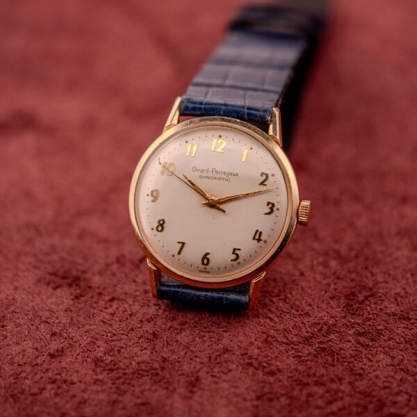 A Girard Perregaux Gyromatic with a white dial, gold Arabic numerals, and a blue strap is set on a textured red surface.