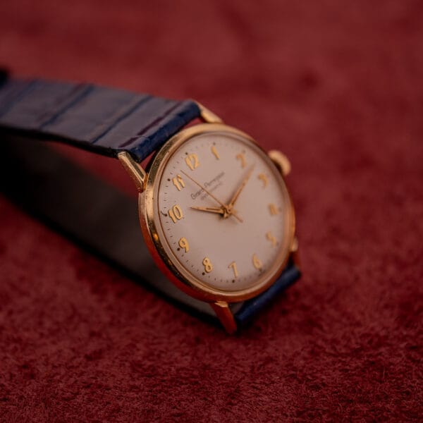 Girard Perregaux Gyromatic with a white dial and gold Arabic numerals, displayed on a blue strap atop a textured burgundy surface.