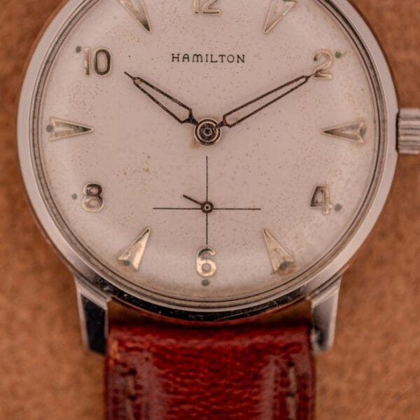 Close-up of a Hamilton Sea-Crest Stainless Steel Case & White Dial Cal. 673 wristwatch with a brown leather strap, shown on a tan background.