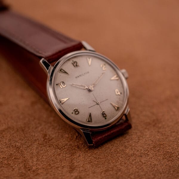 A Hamilton Sea-Crest Stainless Steel Case & White Dial Cal. 673 wristwatch with a brown leather strap rests elegantly on a textured tan surface.