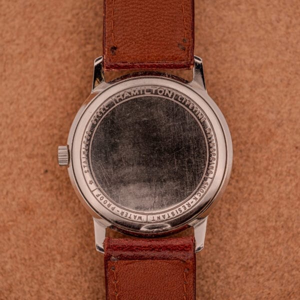 A Hamilton Sea-Crest Stainless Steel Case & White Dial Cal. 673 with a brown leather strap, engraved case back, displayed on a tan textured surface.