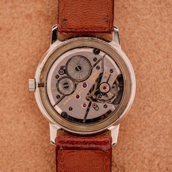 A close-up of the Hamilton Sea-Crest Stainless Steel Case & White Dial Cal. 673 shows its exposed mechanical movement, brown leather strap, and white dial on a tan surface.