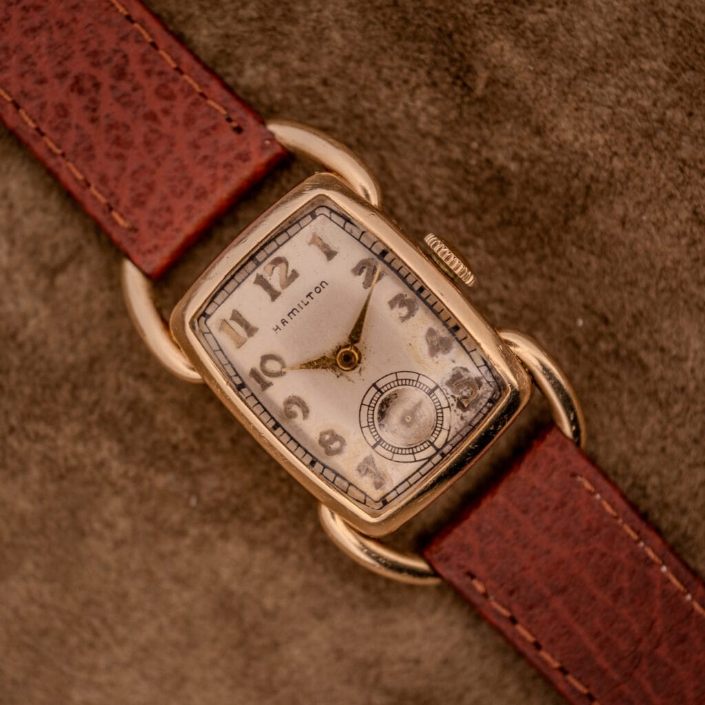 A Hamilton Bowman Art Deco 10k Gold Filled Case Manual Wind Cal. 980 wristwatch with a brown leather strap rests on a textured surface, highlighting its vintage charm and classic style.
