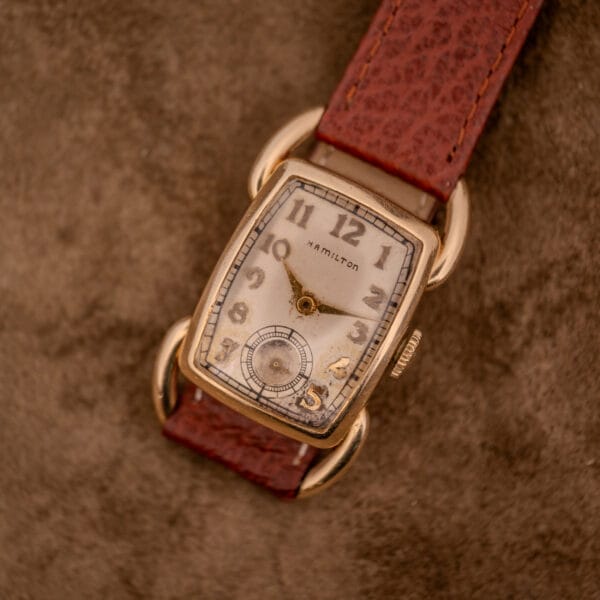 A Hamilton Bowman Art Deco 10k Gold Filled Case Manual Wind Cal. 980 wristwatch with a brown leather strap rests on suede, highlighting its timeless elegance.