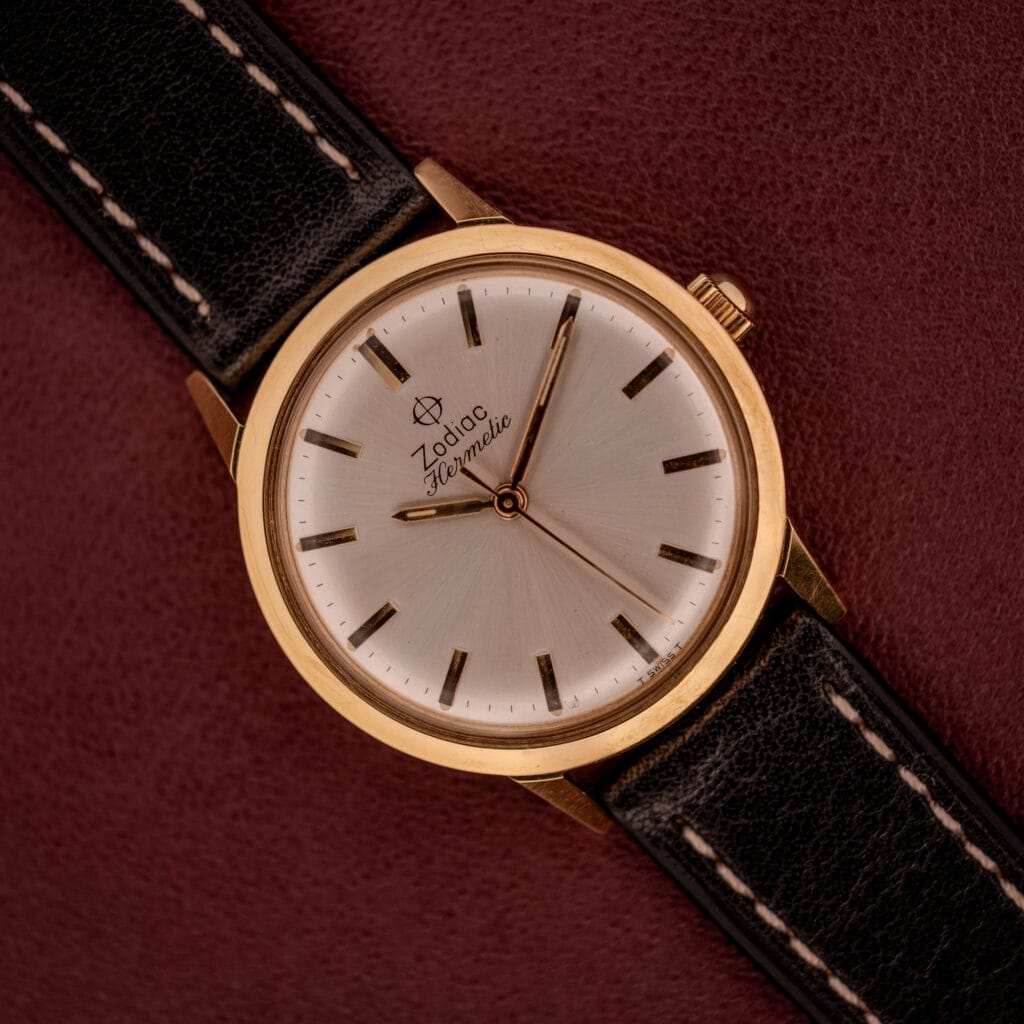 The Zodiac Hermetic Ref. 673 853 Sunburst Dial with a black leather strap rests on a maroon textured surface, shown with its original box and papers.
