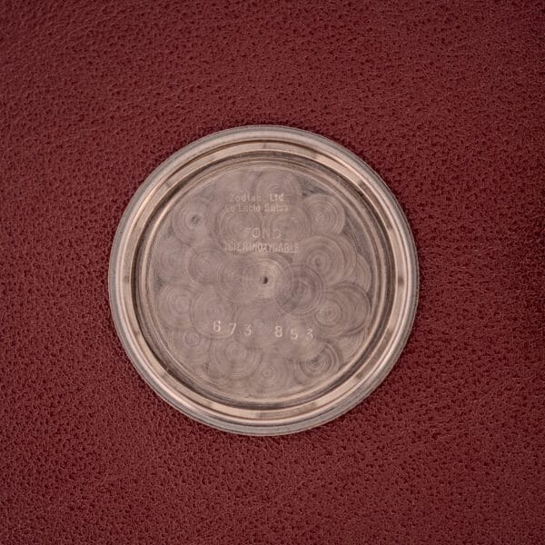 A round metal watch case back with engraved text and numbers rests on a textured reddish-brown surface—an ideal match for the Zodiac Hermetic Ref. 673 853 Sunburst Dial Original Box & Papers.