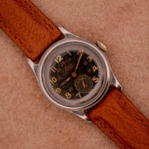 Mido Multifort Super-Automatic Bumper wristwatch with a black and red military dial, gold numerals and hands, paired with a brown leather strap on a tan surface.