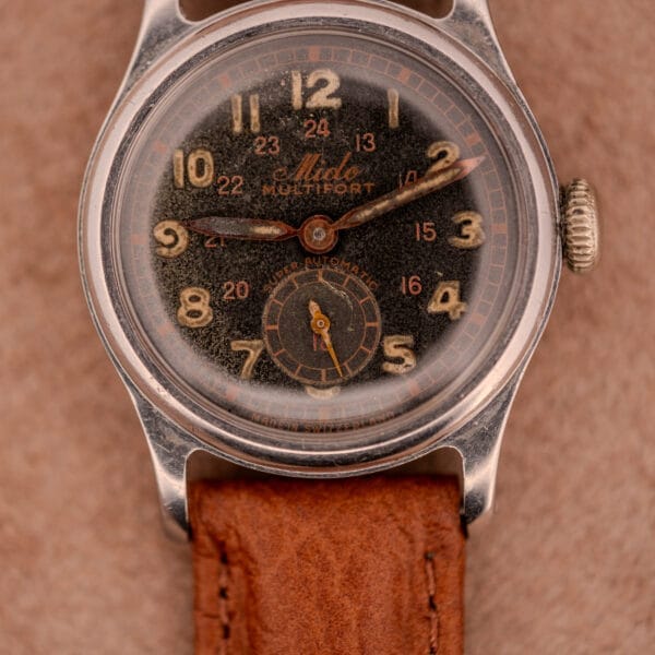 Vintage Mido Multifort Super-Automatic Bumper wristwatch with a black and red military dial, shown on a brown leather strap against a tan background.