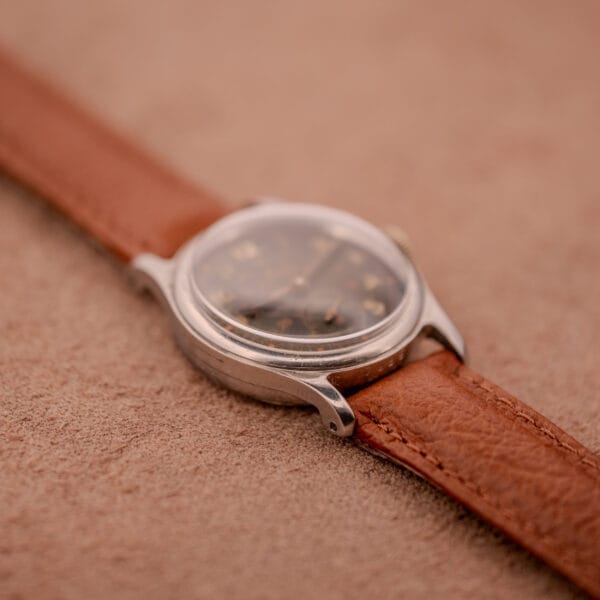 The Mido Multifort Super-Automatic Bumper with a black & red military dial and brown leather strap rests on a soft beige surface.