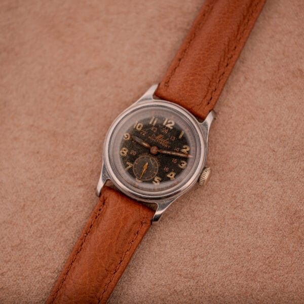 The Mido Multifort Super-Automatic Bumper with a black & red Military Dial and brown leather strap rests on a tan suede background.