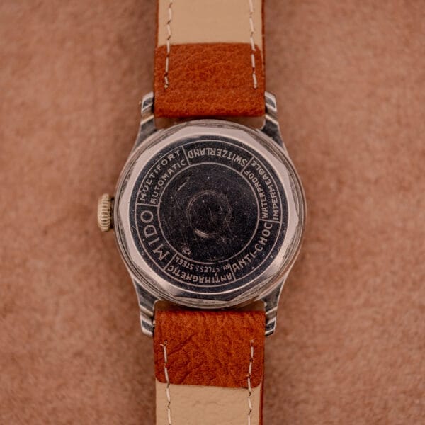 Back view of a vintage Mido Multifort Super-Automatic Bumper Black & Red Military Dial watch with brown leather strap on a light brown surface.