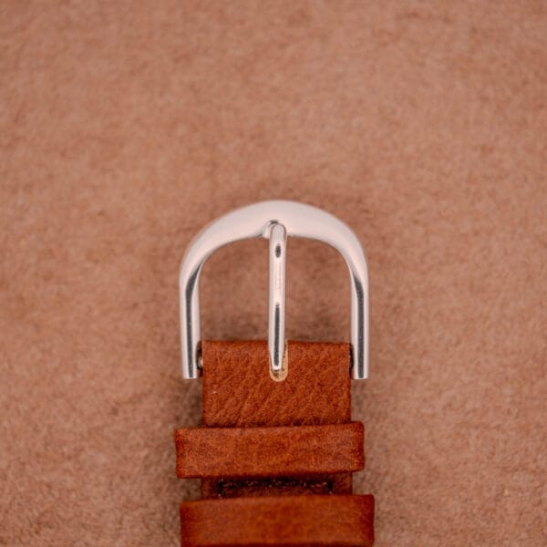 A close-up of a brown leather belt with a silver buckle on a light brown textured background, evoking the rugged style of the Mido Multifort Super-Automatic Bumper Black & Red Military Dial watch.