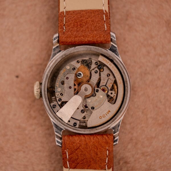 A close-up of the Mido Multifort Super-Automatic Bumper with a black & red military dial and brown leather strap, showcasing its visible mechanical movement.