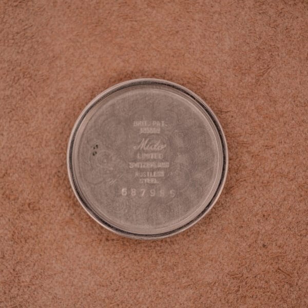 A round metal case back engraved with "Mido Multifort" from the Mido Multifort Super-Automatic Bumper Black & Red Military Dial rests on a textured brown surface.