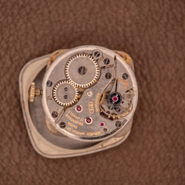 Close-up of the Gruen Veri-Thin Ref. 415-810 with Art Deco lugs, showcasing its exposed manual wind Cal. 415 mechanism and visible gears on a brown textured background.